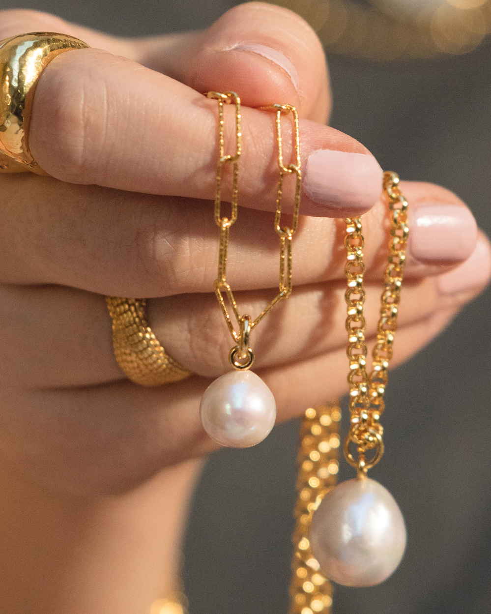 Close up of a model's hand holding a few Nura Baroque Pearls, styled on a variety of gold chains. 